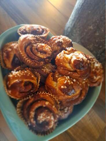 A bowl is filled with golden brown cinnamon buns topped with pearl sugar, in paper liners. The background features a wooden table, creating a cozy atmosphere.
