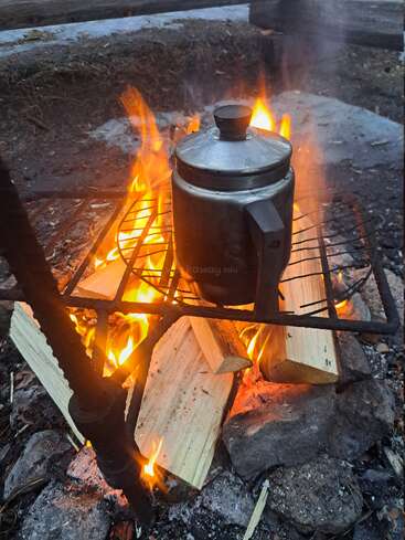 A metal coffee pot sits on a grill above a campfire, surrounded by flaming logs and stones, outdoors, with smoke and rustic ground visible.