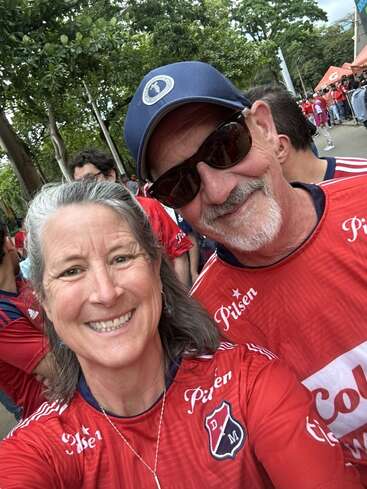 Un homme et une femme souriants prennent un selfie en plein air, tous deux vêtus de maillots rouges de l'équipe de football de l'Independiente Medellín, entourés de gens et d'arbres, profitant d'un événement animé.