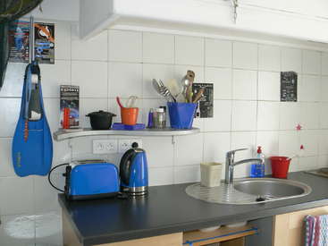 The image depicts a kitchen with white tiles, a black countertop, and various appliances, including a toaster, kettle, and sink, with a few utensils and cleaning supplies nearby.