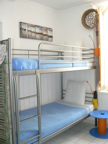 The image depicts a compact room featuring a bunk bed with blue blankets and white pillows, accompanied by a small table and a window with a fishing net hanging on the wall.