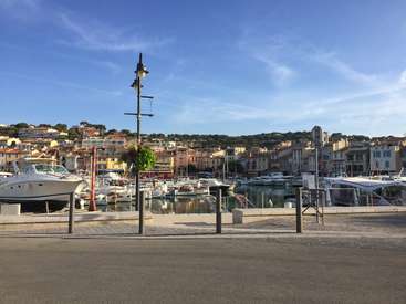 The image depicts a serene marina scene, featuring a row of boats docked along the water\'s edge, with a picturesque town in the background under a clear blue sky.