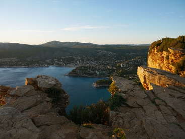 The image depicts a serene coastal town nestled between mountains, with a large body of water and a rocky cliffside in the foreground, bathed in warm sunlight.