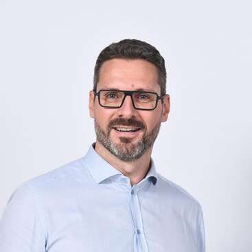 Un homme souriant aux cheveux bruns courts, portant une barbe et des lunettes, vêtu d'une chemise à boutons bleu clair, se tient debout sur un fond clair uni, l'air amical.