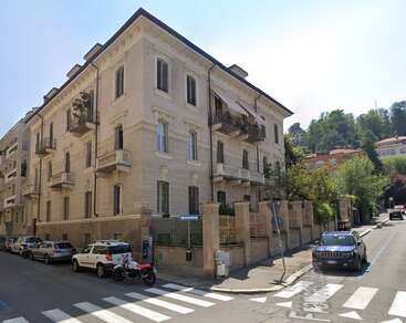 Immeuble d'angle de trois étages avec balcons, façade crème et détails ornementaux. Des voitures et des motos sont garées le long de la rue. Des arbres et de la verdure entourent la zone.