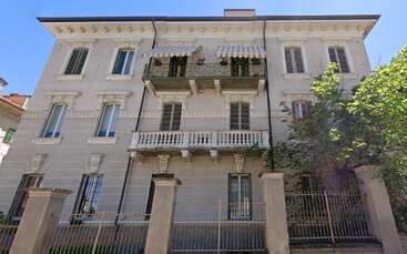 Cette image montre un élégant bâtiment de trois étages avec des balcons ornés, de hautes fenêtres, des moulures décoratives, des grilles en fer forgé et un arbre ombrageant partiellement le côté droit.