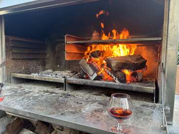 Uma churrasqueira de metal tem lenha queimando com chamas brilhantes. Abaixo, uma taça de vinho tinto está na borda, dando uma vibe relaxada e aconchegante.