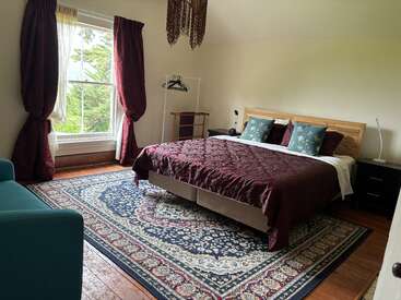 A cozy bedroom with a large bed, burgundy bedding, dark curtains, a blue carpet, teal sofa, wooden floor, large window, and minimal modern decor.