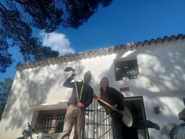 Dos personas posan alegres delante de una casa blanca, con herramientas de jardinería en las manos. El cielo azul y las sombras de los árboles adornan la escena, creando un ambiente rural luminoso y alegre.
