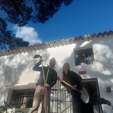 Duas pessoas estão sorridentes em frente a uma casa branca, segurando ferramentas de jardinagem. O céu azul e as sombras das árvores embelezam a cena, criando uma atmosfera rural alegre e luminosa.