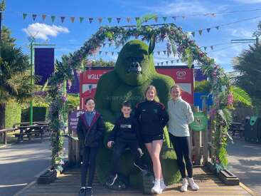 Cuatro niños posan delante de una gran estatua topiaria de un gorila verde. La estatua está enmarcada por un arco floral decorativo en un día soleado.