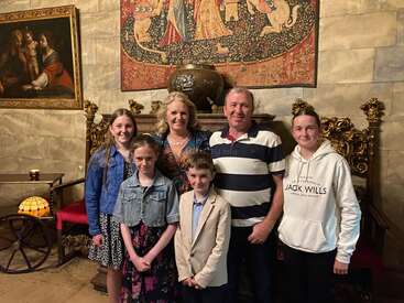 Una familia de seis miembros posa sonriente en una sala adornada con sillas doradas, tapices de estilo medieval y pinturas clásicas en paredes de piedra, que desprenden calidez y unión.