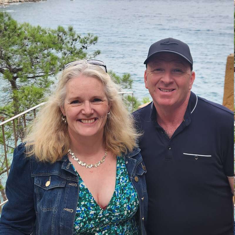 Una pareja sonriente posa al aire libre junto al agua, con árboles y una costa rocosa de fondo. La mujer lleva un vestido estampado y una cazadora vaquera; el hombre, una gorra.