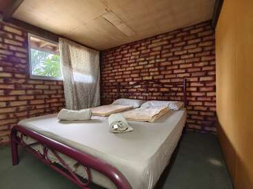 Una acogedora habitación con paredes de ladrillo cuenta con una cama doble, cabecero metálico, toallas dobladas, cortinas sencillas y una ventana que deja entrar la luz natural, creando un ambiente rústico.
