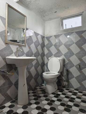 Esta imagen muestra un cuarto de baño pequeño con azulejos de motivos geométricos, un lavabo con pedestal, un espejo de pared, un inodoro con la tapa abierta, un bidé de mano y una ventana pequeña.