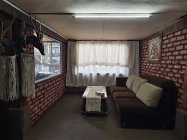 Esta acogedora habitación cuenta con un sofá marrón, una mesa de centro, paredes de ladrillo, una gran ventana con cortinas, cabezas de antílope disecadas, decoración de macramé y un retrato familiar.