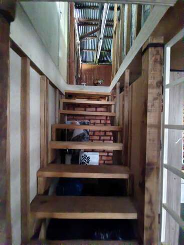 Una estrecha escalera de madera asciende entre vigas de madera sin terminar, conduciendo a un piso superior con techo de metal corrugado. Se pueden ver vigas expuestas y diversos objetos.