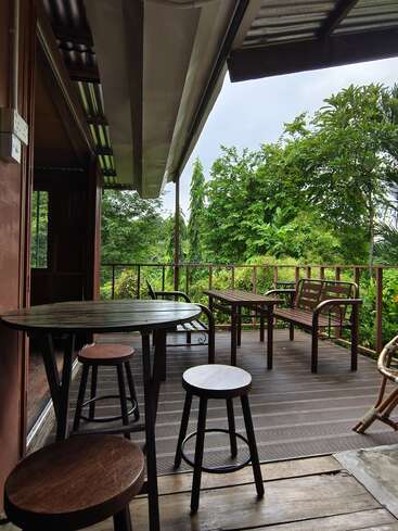Un acogedor patio de madera con taburetes y un banco ofrece vistas a frondosos árboles verdes. La terraza cubierta proporciona un lugar tranquilo y relajante para disfrutar de la belleza de la naturaleza exterior.