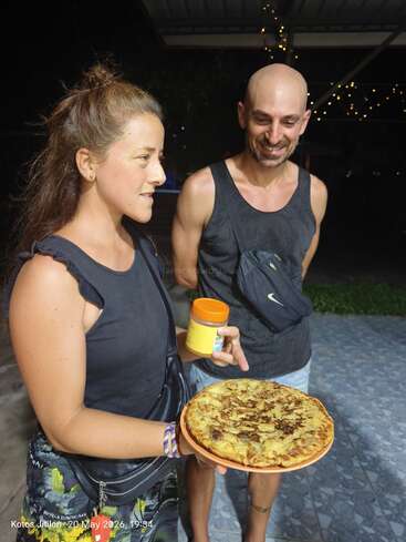 A woman holds a round, golden-brown omelette on a plate and a jar, while a smiling man stands beside her. String lights twinkle in the background.