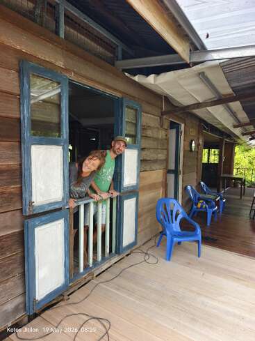 A man and woman lean playfully out of a rustic wooden window. Blue chairs sit on a porch. Lush greenery surrounds the cozy, rural house setting.