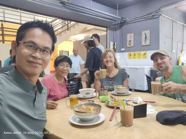 Four smiling people sit around a table enjoying food and drinks at a casual indoor eatery. The atmosphere is cheerful, with more diners and kitchen staff visible.