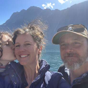 A smiling family stands together outdoors by a lake, with mountains in the background. One person kisses another’s cheek, capturing a joyful, sunny moment.