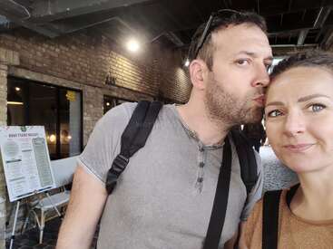 The image shows a man kissing a woman on the forehead, both wearing backpacks, with a brick wall and ceiling visible in the background.