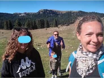 The image shows a family of four posing for a selfie in a grassy field with trees and mountains in the background on a sunny day.