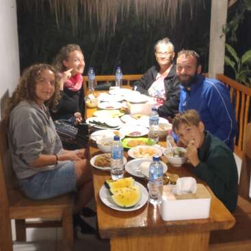 Sechs Leute sitzen um einen Holztisch herum und essen zusammen unter einer Hängelampe. Der Tisch ist voll mit Essen, Wasserflaschen und Obst.