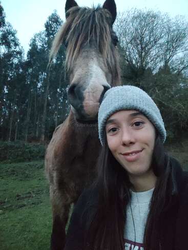 Uma jovem mulher, usando um gorro cinza, sorri enquanto tira uma selfie com um grande cavalo marrom parado logo atrás dela em uma área verde e arborizada.