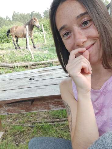 Uma jovem sorridente tira uma selfie ao ar livre, sentada em uma mesa de madeira. Atrás dela, um cavalo marrom está em uma área gramada cercada com árvores.