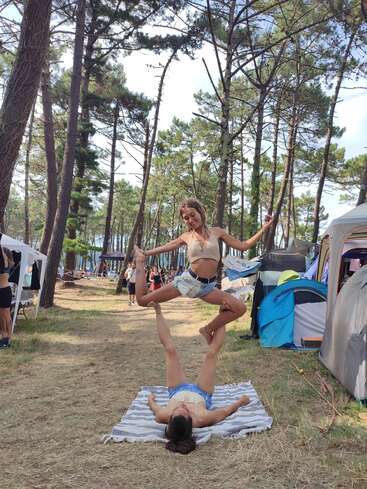 Duas mulheres estão praticando acroioga em um tapete listrado em um acampamento na floresta, cercado por árvores altas e tendas coloridas, com pessoas observando nas proximidades.
