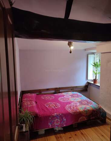 Esse quarto aconchegante tem uma cama baixa com uma colcha rosa com padrão de mandala colorida, piso de madeira, viga preta exposta no teto, uma pequena planta e uma janela aberta.