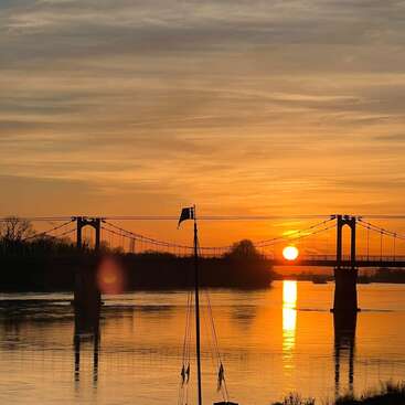 A imagem mostra um pôr do sol dourado sobre um rio calmo, com uma ponte suspensa, árvores em silhueta e o mastro de um veleiro refletido na água cintilante.