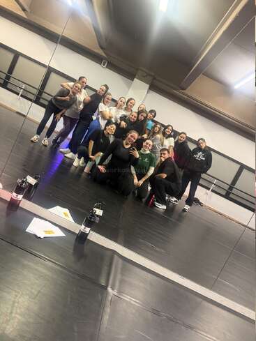 Un groupe de personnes pose ensemble dans un studio de danse, souriant devant un miroir mural. Des bouteilles d'eau et des papiers se reflètent sur le sol.