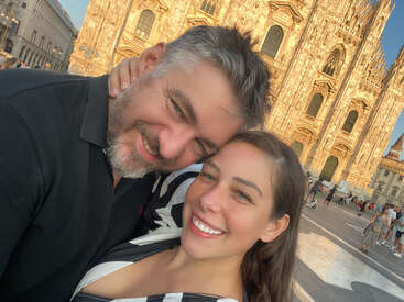 Un couple heureux prend un selfie en gros plan devant une cathédrale historique baignée de soleil. Ils sourient chaleureusement, profitant ensemble de leur voyage sur une place animée.