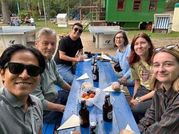 Six people sit around a blue picnic table outdoors, smiling for a group selfie. Food, drinks, and table tennis tables are visible, suggesting a casual gathering.