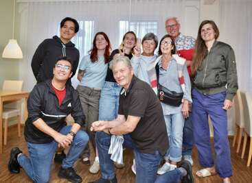 A group of nine people of various ages pose cheerfully indoors. Some are standing, some kneeling, all smiling. The atmosphere is casual and friendly.