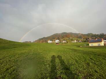 A green field stretches towards charming houses beneath a forested hill. A vivid rainbow arcs across a cloudy sky. Two shadows stand together in the foreground.