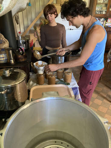 Two people are canning food in a cozy kitchen. One ladles mixture into jars while the other holds a spoon. Pots and canning equipment are visible.