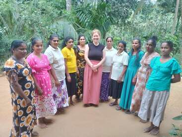 Un grupo de once mujeres permanecen juntas al aire libre sobre un suelo arenoso, rodeadas de frondosas plantas y árboles, todas sonrientes y vestidas con ropas de colores.