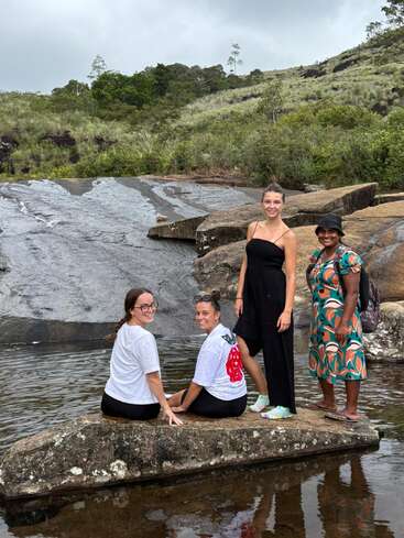 Four women enjoy a serene natural landscape, sitting and standing on rocks by a stream, surrounded by lush greenery, hills, and overcast skies, smiling together.