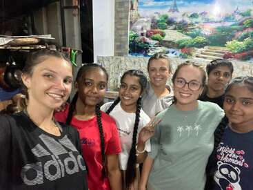A group of seven young women stand together, smiling at the camera. They appear happy and relaxed, posing indoors with a colorful mural in the background.