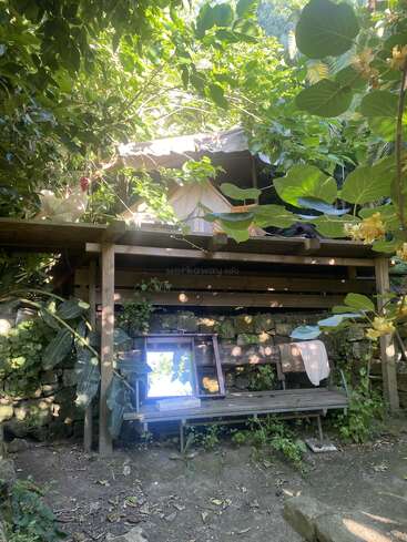 A cozy tent sits atop a wooden platform surrounded by lush green foliage. Beneath it, a bench, mirror, and some items create a tranquil, rustic hideaway.
