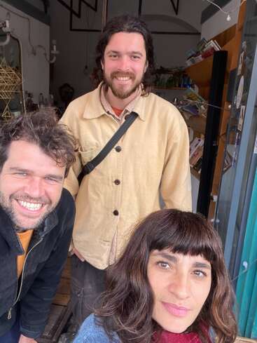 Three people are smiling warmly at the camera, standing closely together in a cozy indoor setting with shelves and decorative items in the background. Friendly atmosphere.