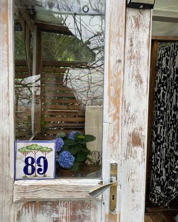 Uma porta de madeira branca desgastada pelo tempo, com um painel de vidro, revela hortênsias azuis no interior. O número 89, feito de cerâmica, está à vista, com reflexos naturais visíveis no vidro.