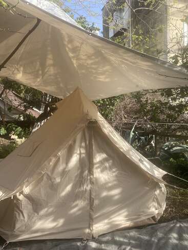 Uma barraca de lona bege está montada sob uma grande lona, num jardim exuberante. A luz do sol penetra, projetando sombras suaves sobre o tecido.