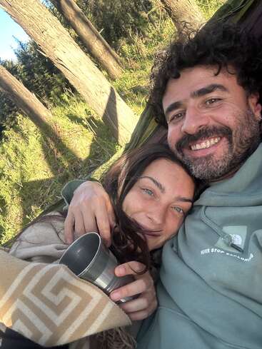 Das Bild zeigt einen Mann und eine Frau in einer Hängematte, wobei die Frau eine Metalltasse in der Hand hält, vor einem Hintergrund aus Bäumen und Gras an einem sonnigen Tag.