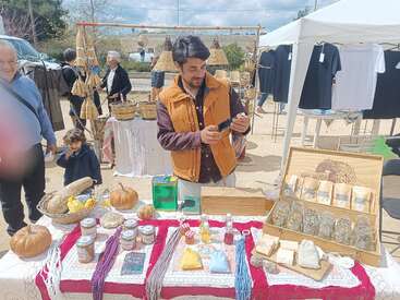 Ein Mann steht an einem Marktstand im Freien und verkauft Gläser, Kräuter, Seifen und handgemachtes Kunsthandwerk. Andere Leute stöbern an den Ständen in der sonnigen, lebhaften Umgebung.