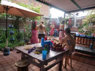A group of people gathers in a cozy outdoor patio with plants, a covered roof, and a table set with cups, utensils, and food for a celebration.